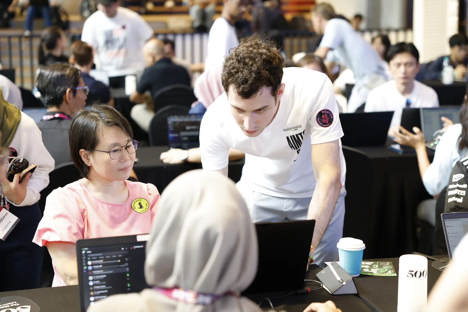 A Malaysian AI volunteer kneeling beside a participant and helping him work through his project